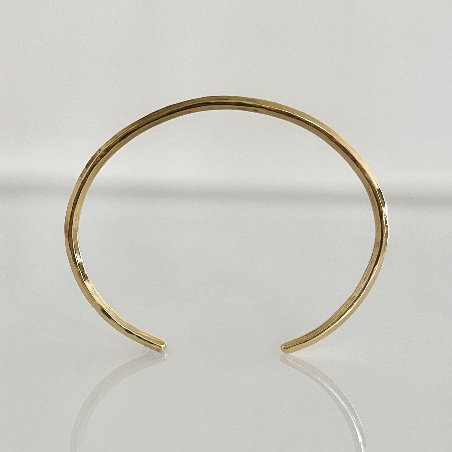 An open cuff bracelet made of 14k solid gold with a raw, hammered finish, displayed on a reflective surface.