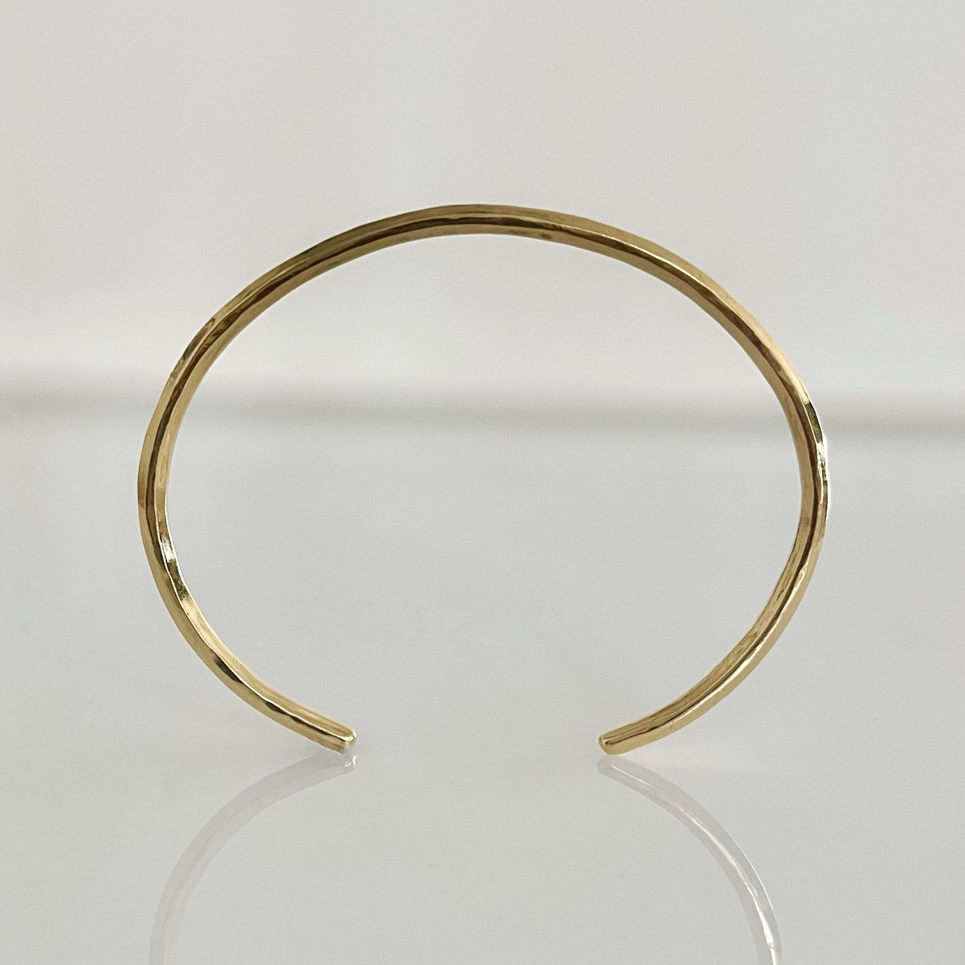 An open cuff bracelet made of 14k solid gold with a raw, hammered finish, displayed on a reflective surface.