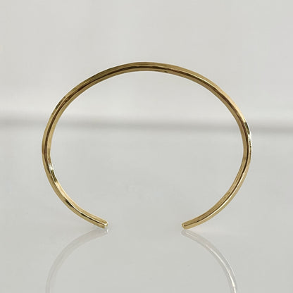 An open cuff bracelet made of 14k solid gold with a raw, hammered finish, displayed on a reflective surface.