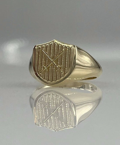 A 14K solid gold shield-shaped signet ring with a design of crossed swords on a textured crest, displayed against a reflective background.